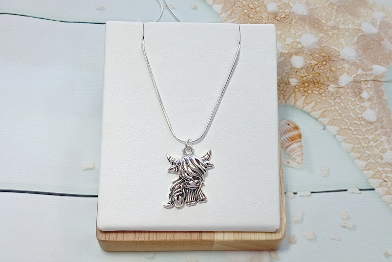 May include: A silver-toned necklace featuring a pendant shaped like a Highland cow with long, shaggy hair and curved horns. The pendant hangs from a thin, smooth silver-toned chain, creating a detailed and metallic accessory for everyday wear.