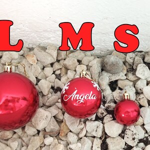May include: Three red Christmas ornaments of varying sizes, resting on a bed of small white stones. The largest ornament is plain, the middle one has the name "Angela" and snowflake designs, and the smallest is plain. The letters "LMS" are above.