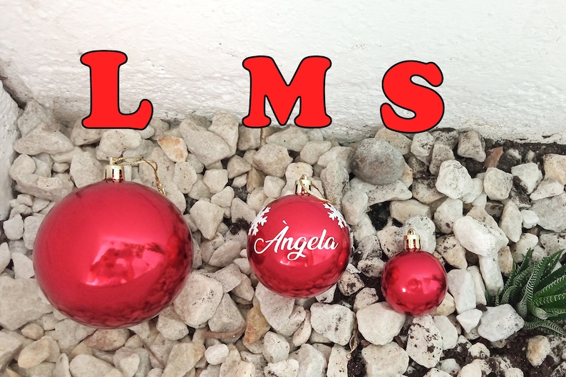 May include: Three red Christmas ornaments of varying sizes, resting on a bed of small white stones. The largest ornament is plain, the middle one has the name "Angela" and snowflake designs, and the smallest is plain. The letters "LMS" are above.
