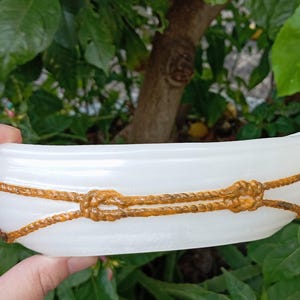 Op de afbeelding: Een witte, bootvormige decoratieve schaal met een goudkleurig touwontwerp. Het touw is in knopen om de schaal geknoopt. De schaal wordt in een hand gehouden tegen een achtergrond van groene bladeren en een boomstam.