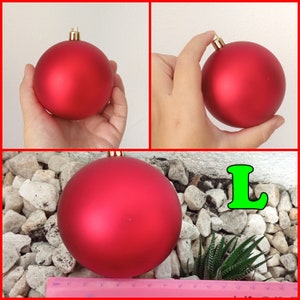 May include: A large, matte red Christmas ornament with a gold cap. The ornament is held in a hand in two of the images. The third image shows the ornament on a bed of rocks with a small succulent plant and a ruler.