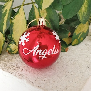 May include: A shiny red Christmas ornament with the name "Angela" in white script, accented by white snowflake designs. The ornament hangs from a gold string, perfect for holiday decorations.
