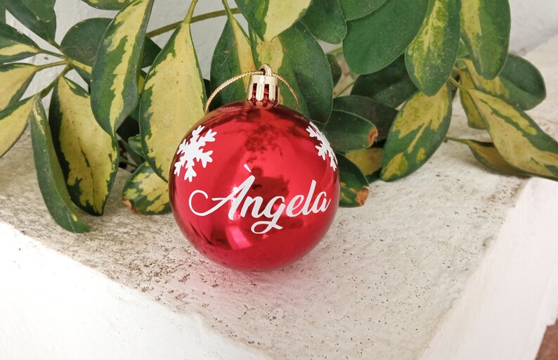 May include: A shiny red Christmas ornament with the name "Angela" in white script, accented by white snowflake designs. The ornament hangs from a gold string, perfect for holiday decorations.