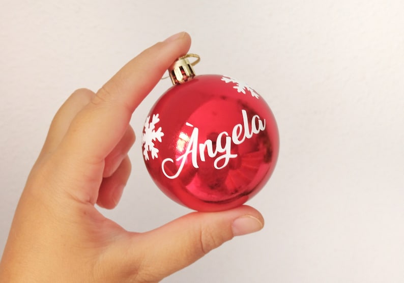 May include: A shiny red Christmas ornament with the name "Angela" in white script. White snowflake designs are also on the ornament. The ornament has a gold-colored metal cap.