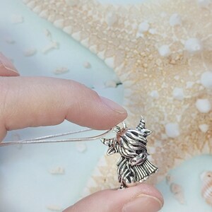 May include: A silver-toned metal pendant shaped like a Highland cow with long, shaggy hair, curved horns, and a detailed face, attached to a thin, flexible silver-colored chain necklace that rests against the pendant's top loop.