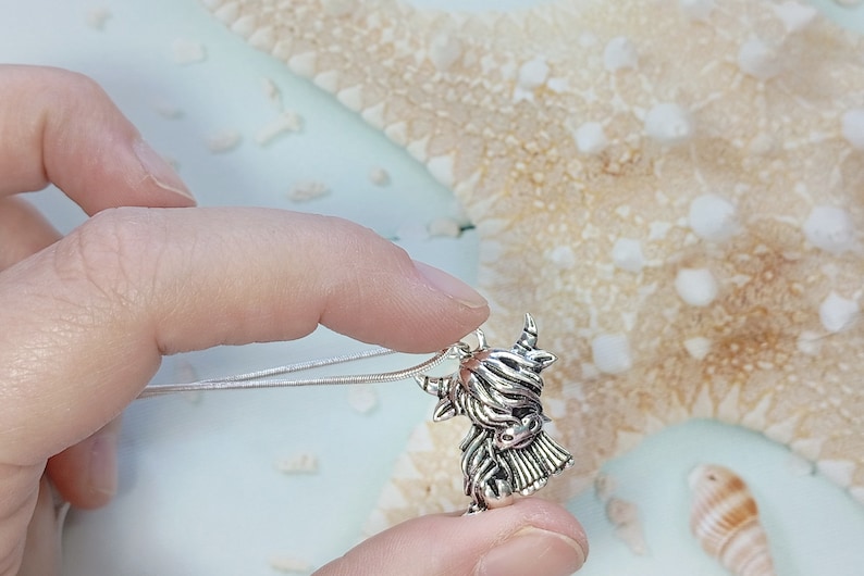 May include: A silver-toned metal pendant shaped like a Highland cow with long, shaggy hair, curved horns, and a detailed face, attached to a thin, flexible silver-colored chain necklace that rests against the pendant's top loop.