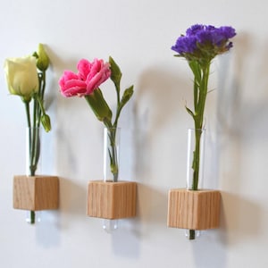 Op de afbeelding: Drie houten wandvasen met glazen reageerbuizen. De vazen zijn gemaakt van natuurlijk hout en hebben een lichtbruine kleur. De reageerbuizen zijn van helder glas en bevatten elk een enkele witte, roze en paarse bloem.