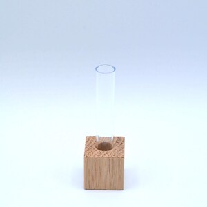May include: A small, clear glass test tube vase sits in a square wooden holder. The wood is a light brown color.