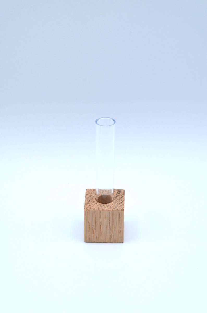 May include: A small, clear glass test tube vase sits in a square wooden holder. The wood is a light brown color.