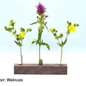 May include: A wooden flower holder with three glass tubes, holding a purple flower and two yellow flowers. The holder is labeled "15cm Halter: Walnuss".