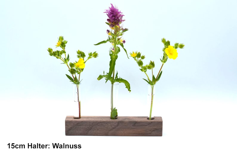May include: A wooden flower holder with three glass tubes, holding a purple flower and two yellow flowers. The holder is labeled "15cm Halter: Walnuss".