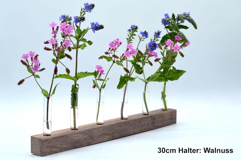 May include: A wooden flower holder with five glass tubes, each holding a different type of flower. The flowers are pink and blue. The holder is 30cm long and made of walnut wood.