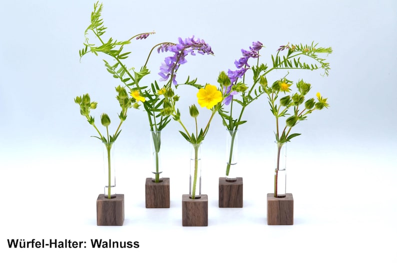 May include: Six small wooden cube holders with glass test tubes filled with wildflowers. The flowers are purple, yellow, and green. The text "Wuerfel-Halter: Walnuss" is printed below the holders.