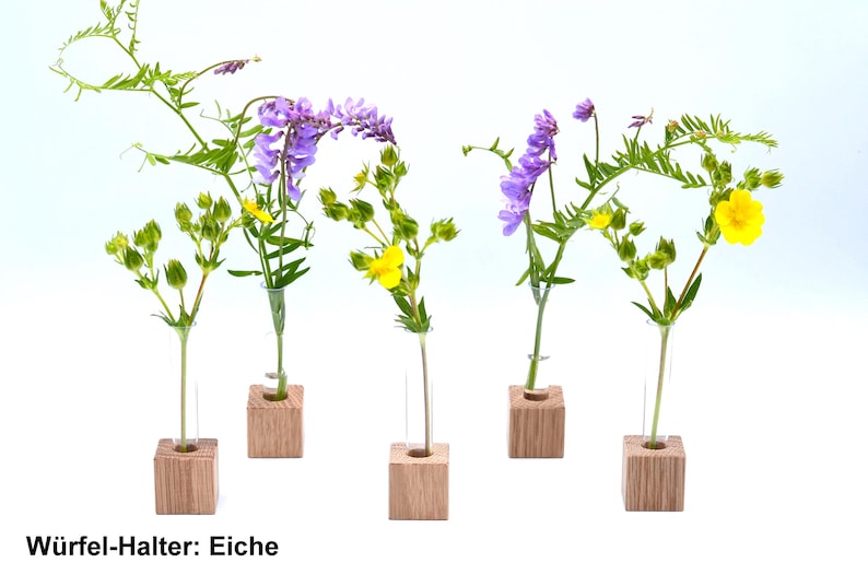 May include: Five small wooden cube holders with glass test tubes, each holding a different type of flower. The flowers are purple, yellow, and green. The text "Wuerfel-Halter: Eiche" is printed below the holders.