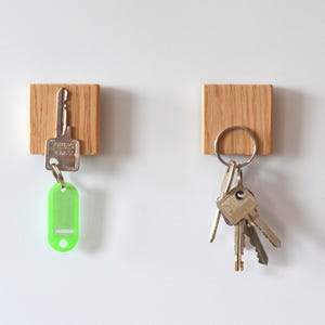 Op de afbeelding: Twee houten sleutelhangers aan een witte muur. Eén houdt een enkele sleutel met een groen label vast, de andere houdt een set sleutels aan een metalen ring vast. De sleutelhangers zijn vierkant en gemaakt van lichtgekleurd hout.