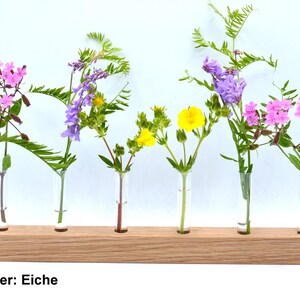 May include: A wooden flower holder with six glass tubes, each holding a different type of flower. The flowers are pink, purple, and yellow. The holder is labeled "30cm Halter: Eiche".