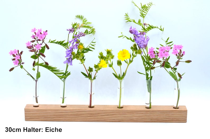 May include: A wooden flower holder with six glass tubes, each holding a different type of flower. The flowers are pink, purple, and yellow. The holder is labeled "30cm Halter: Eiche".