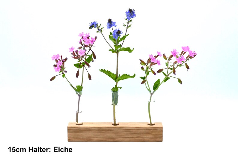 May include: Three glass bud vases with a wooden holder. The vases hold pink and blue flowers. The holder is labeled "15cm Halter: Eiche".
