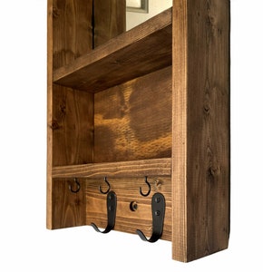 May include: A wooden wall-mounted shelf unit with a dark stain finish. It has two shelves and three black metal hooks. The wood grain is visible, enhancing the natural look. Designed for storage and organisation, it's a practical home accessory.