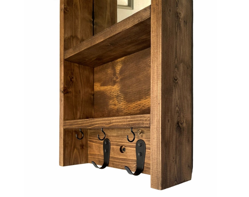 May include: A wooden wall-mounted shelf unit with a dark stain finish. It has two shelves and three black metal hooks. The wood grain is visible, enhancing the natural look. Designed for storage and organisation, it's a practical home accessory.