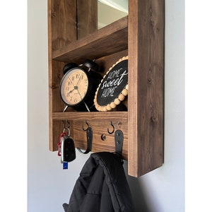 May include: A dark stained wooden wall shelf unit. It has a top shelf with a mirror, a middle shelf with a black alarm clock and a sign reading "Home Sweet Home", and a lower shelf with hooks for keys and a coat.