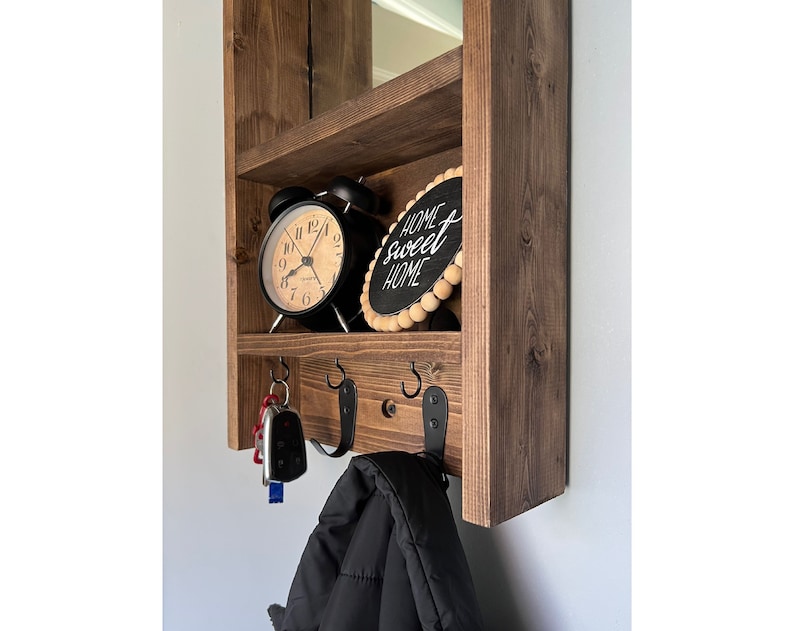 May include: A dark stained wooden wall shelf unit. It has a top shelf with a mirror, a middle shelf with a black alarm clock and a sign reading "Home Sweet Home", and a lower shelf with hooks for keys and a coat.