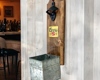Op de afbeelding: Een aan de muur gemonteerde flesopener met een donkere houten achterkant en een metalen flesopener. Een geel bord met het "Coors Banquet" logo is aan het hout bevestigd. Een gegalvaniseerde metalen container bevindt zich eronder.