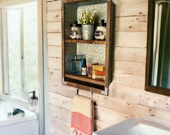 May include: A rustic wooden bathroom shelf with a galvanized metal backing. The shelf holds jars of cotton balls, a small plant, soaps, and a blue perfume bottle. A striped towel hangs below.