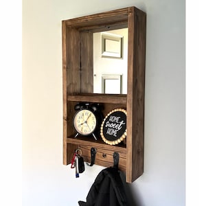 May include: A dark stained wooden wall organiser with a mirror, shelf, and hooks. It includes a black alarm clock, a sign reading "home sweet home", and keys hanging from the hooks. The unit is designed for entryway or hallway use.