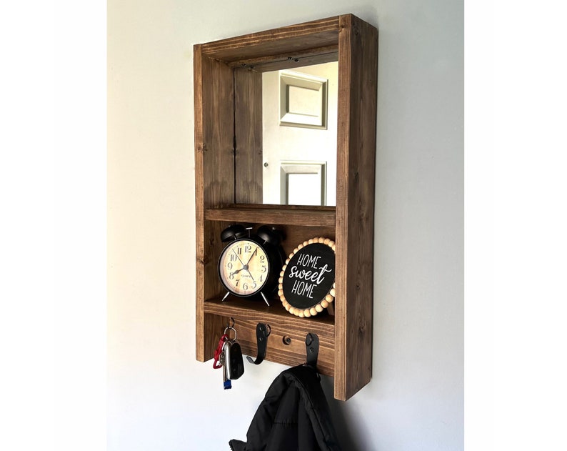 May include: A dark stained wooden wall organiser with a mirror, shelf, and hooks. It includes a black alarm clock, a sign reading "home sweet home", and keys hanging from the hooks. The unit is designed for entryway or hallway use.
