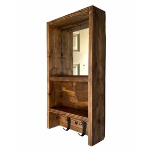 May include: A wooden wall shelf unit with a mirror, two shelves, and three black metal hooks. The wood is stained a warm brown colour. The unit is designed for storage and organisation.