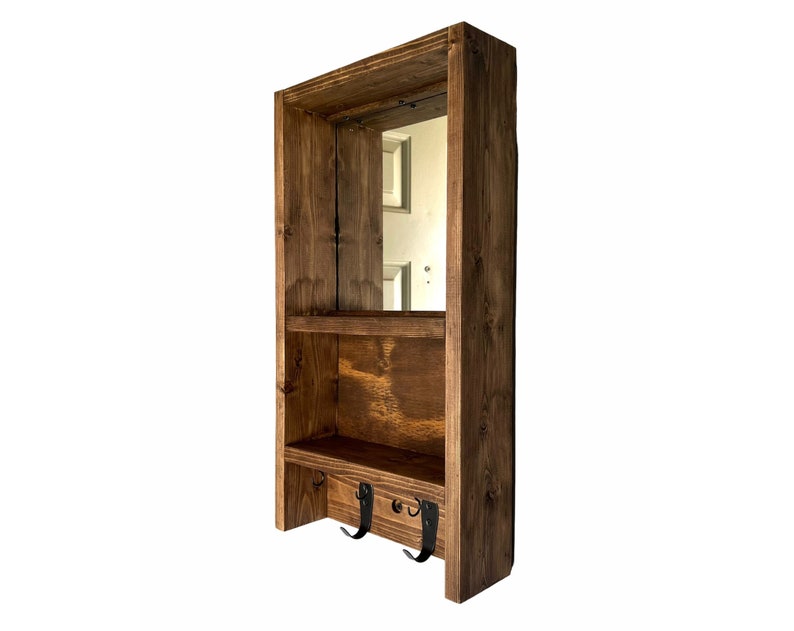 May include: A wooden wall shelf unit with a mirror, two shelves, and three black metal hooks. The wood is stained a warm brown colour. The unit is designed for storage and organisation.