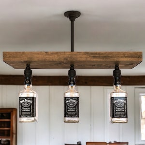May include: A rustic, three-light pendant lamp featuring repurposed Jack Daniel's whiskey bottles as light fixtures. The bottles hang from a dark brown wooden beam, suspended from a ceiling mount. The bottles have black and white labels.
