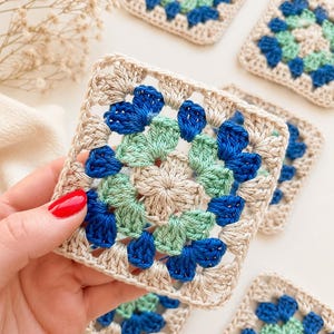 Könnte beinhalten: Handgefertigte Häkeluntersetzer in quadratischer Form. Jeder Untersetzer hat einen beigen Rand mit blauen und hellgrünen Blumenmustern in der Mitte. Die Untersetzer sind aus Garn gefertigt und ideal für die Inneneinrichtung.