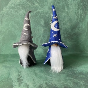 May include: Two felt gnome ornaments with white beards and embroidered hats. The gnome on the left has a gray hat with a white crescent moon and stars. The gnome on the right has a blue hat with a white crescent moon and stars.