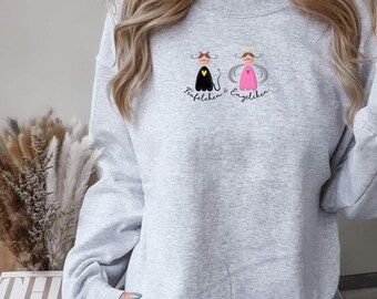Könnte beinhalten: Hellgraues Sweatshirt mit einem Cartoon-Teufel- und Engel-Design und dem Text "Teufelchen & Engelchen". Der Teufel ist schwarz mit einem roten Herz, der Engel ist rosa. Das Sweatshirt hat einen Rundhalsausschnitt.