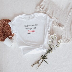 Könnte beinhalten: Weißes T-Shirt mit den Worten "tolerance respect love" in Schwarz und Rot gedruckt. Das Wort "love" hat ein kleines Herzsymbol darunter. Das Shirt ist mit einem geflochtenen Korb, einer weißen Tasse und Blumen gestaltet.