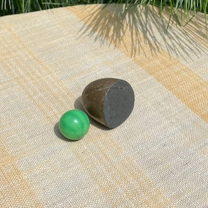 Puede incluir: Un mármol de vidrio verde y un objeto de piedra de color marrón oscuro y negro en forma de huevo sobre una superficie de tela rayada beige y marrón.