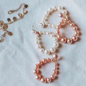 Puede incluir: Tres pares de pendientes de aro de perlas. Los pendientes están hechos de perlas blancas y rosadas. Los pendientes están sobre un fondo de tela blanca.