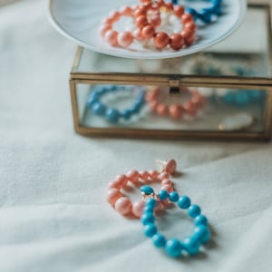 Puede incluir: Dos pares de pendientes de aro, uno rosa y otro azul, sobre un plato blanco y una caja de joyería con espejo. Los pendientes están hechos de cuentas grandes y redondas.