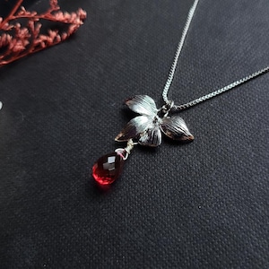 Op de afbeelding: Een zilverkleurige ketting met een metalen bloemhanger met gestructureerde bloemblaadjes. Een gefacetteerde, druppelvormige rode edelsteen hangt in het midden van de bloem. De hanger is bevestigd aan een dunne zilverkleurige ketting. De tekst deahariasa.com is zichtbaar.