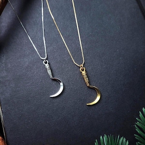 May include: Two necklaces with crescent moon pendants. One necklace is silver, the other is gold. The pendants have detailed handles. The necklaces are on a dark background with a wooden slice and greenery.