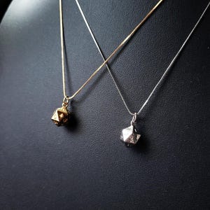 Op de afbeelding: Twee kettingen met een gouden en zilveren geometrische hanger in de vorm van een twintigzijdige dobbelsteen. De kettingen liggen op een zwarte displaystandaard. deahariasasa.com