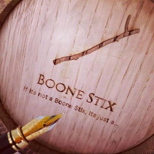 May include: Close-up of a wooden barrel lid with the text "BOONE STIX" and a branch design. A gold-tipped fountain pen is in the foreground. The text "If Its not a Boone Stix, Its just a..." is also visible.