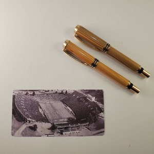 May include: Two gold and wood-toned pens with black accents and a small black and white stadium postcard are displayed on a white surface. The pens have a ribbed design and gold-colored clips.