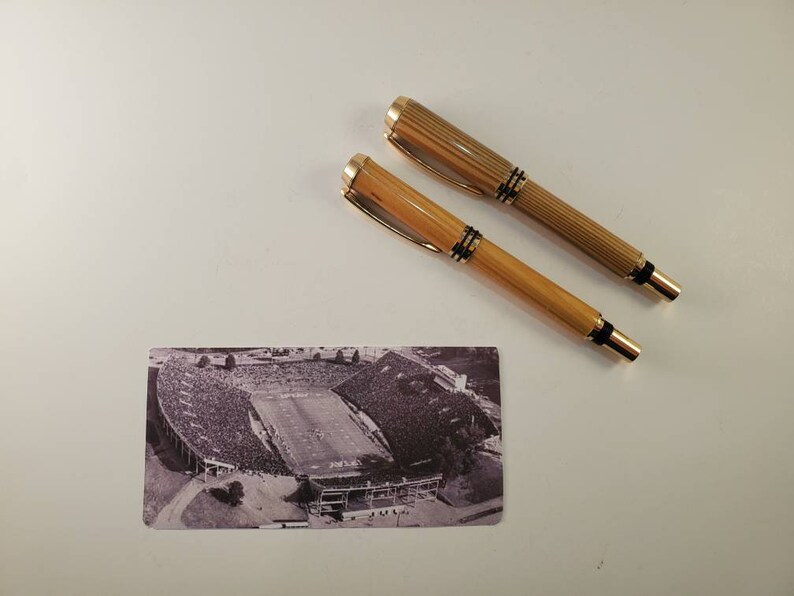 May include: Two gold and wood-toned pens with black accents and a small black and white stadium postcard are displayed on a white surface. The pens have a ribbed design and gold-colored clips.