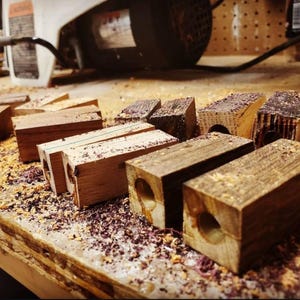 May include: A collection of wooden blocks in various stages of crafting, with a hole drilled through each. The blocks are scattered on a workbench covered in wood shavings, with a blurred background of tools and equipment.