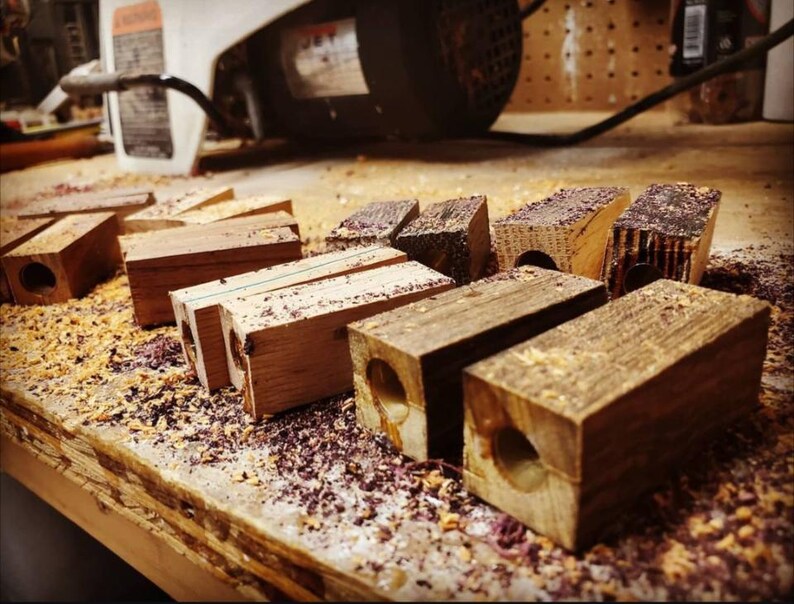 May include: A collection of wooden blocks in various stages of crafting, with a hole drilled through each. The blocks are scattered on a workbench covered in wood shavings, with a blurred background of tools and equipment.