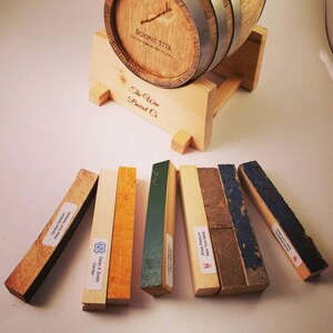 May include: A small wooden barrel with metal bands, labeled "BOONE STIX" and "The Wee Barrel Co." rests on a wooden stand. Several rectangular wooden pieces with labels are arranged in front, displaying various colors and textures.