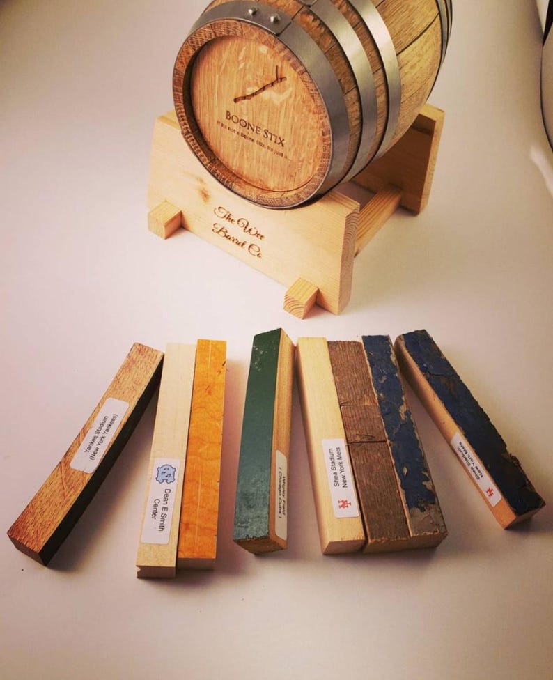 May include: A small wooden barrel with metal bands, labeled "BOONE STIX" and "The Wee Barrel Co." rests on a wooden stand. Several rectangular wooden pieces with labels are arranged in front, displaying various colors and textures.